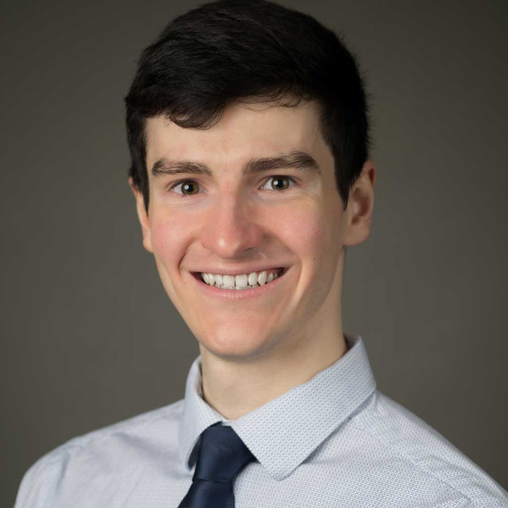 Head-and-shoulders studio portrait of an adult with short dark hair, wearing a light-colored button-down shirt and a dark tie, facing the camera against a plain gray background.